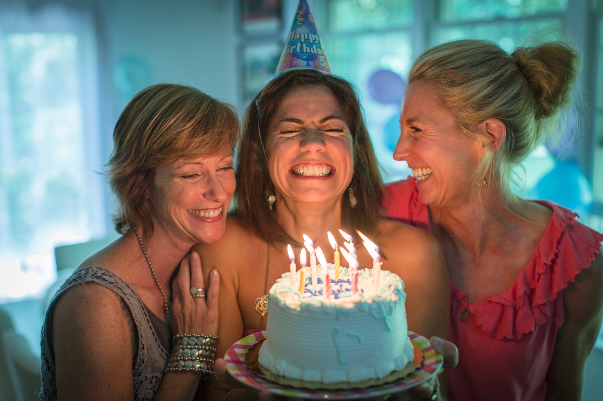 Mature woman holding birthday cake, making wish while two friends look on
