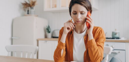 Nervous frowning woman making phone call customer service, sitting at table with notebook in kitchen