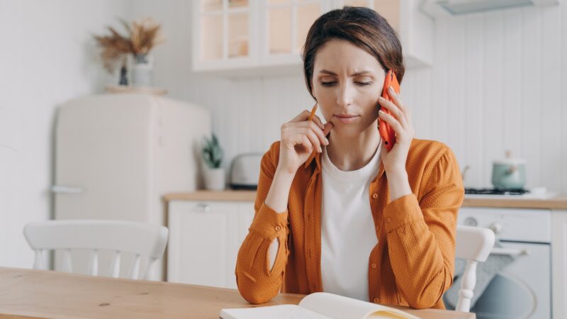 Nervous frowning woman making phone call customer service, sitting at table with notebook in kitchen