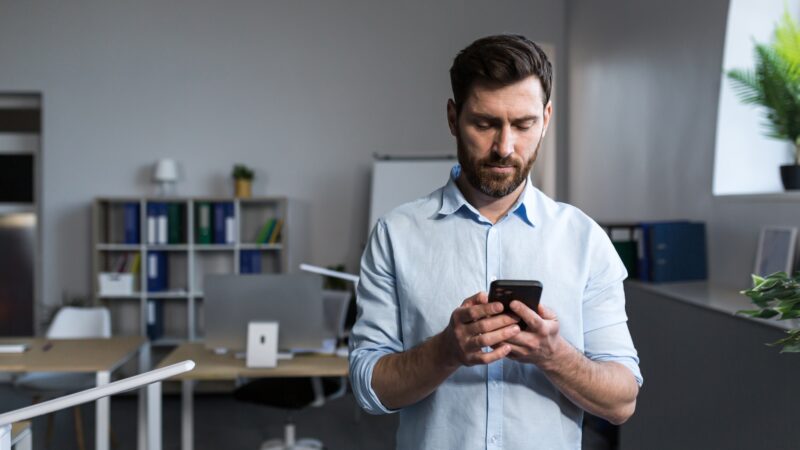 Portrait of a young man in the office. Stands, uses the phone, writes a message