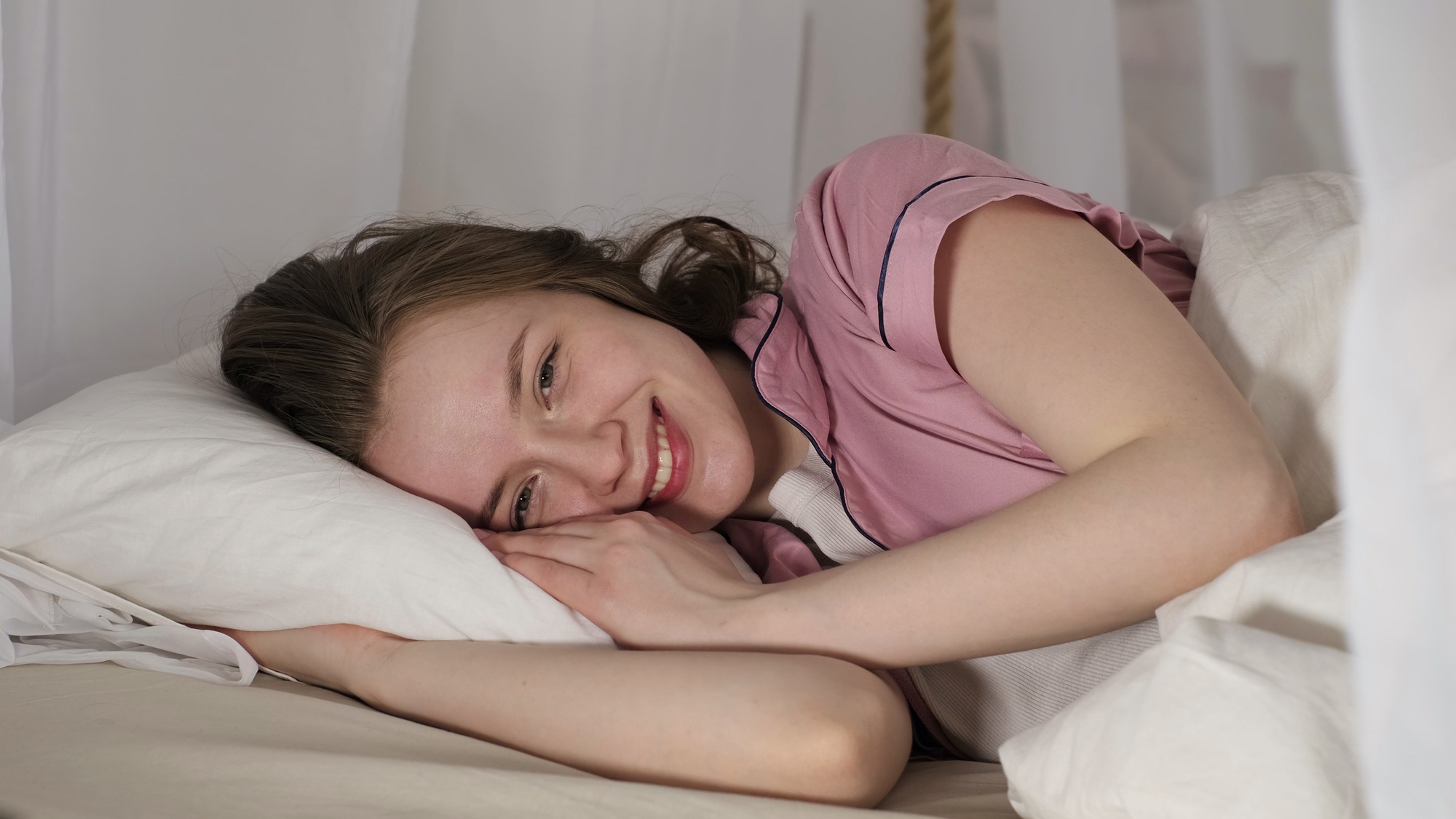 A woman in a pink pajama top smiles while lying on a white pillow, looking content and relaxed. This