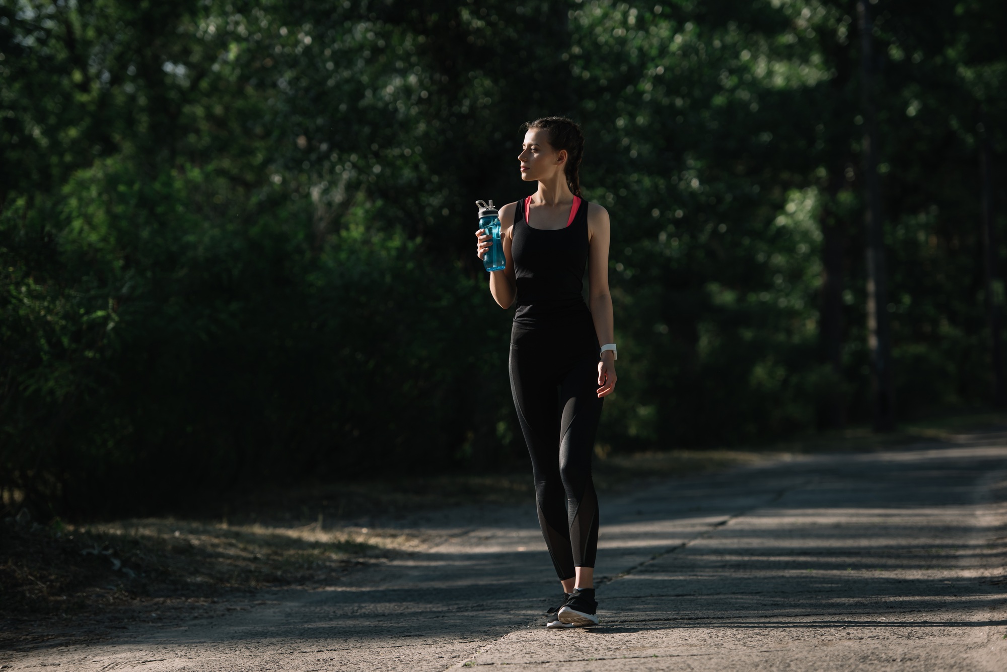 athletic girl holding sports bottle with water and walking in park