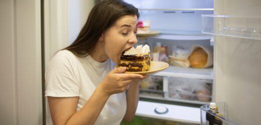 Hungry woman in pajamas eating sweet cakes at night near refrigerator