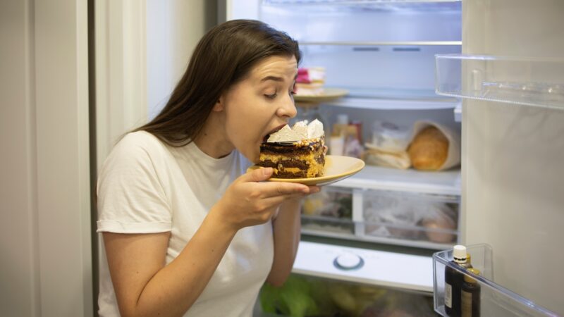 Hungry woman in pajamas eating sweet cakes at night near refrigerator