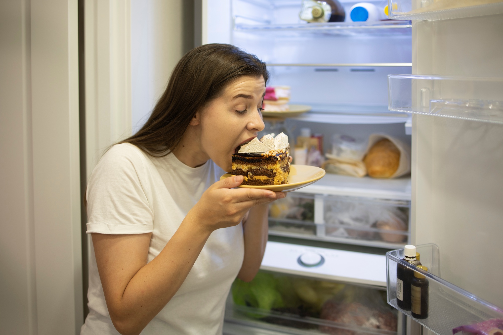Hungry woman in pajamas eating sweet cakes at night near refrigerator